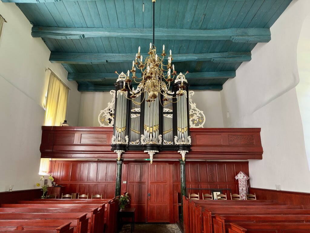 Het interieur van de Laurentiuskerk in Baflo. Een groen, oud, houten plafond. Een orgel, kroonluchter en roodgeschilderde houten banken en ballustrade.