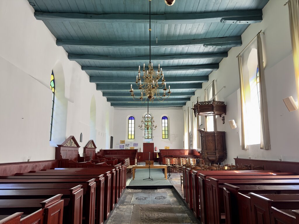 Het interieur van de Laurentiuskerk in Baflo. Een groen, oud, houten plafond. Een kroonluchter en roodgeschilderde houten banken en ballustrade.