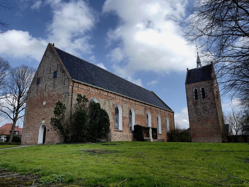 De Laurentiuskerk met toren in Baflo. Omringd door kale bomen, groen gras en een blauwe lucht.
