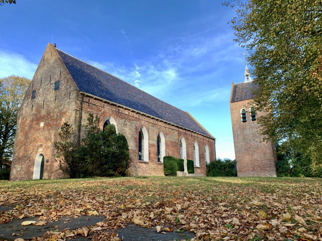 De Laurentiuskerk met toren in Baflo. Omringd door bomen in herfstkleuren, groen gras en een blauwe lucht.