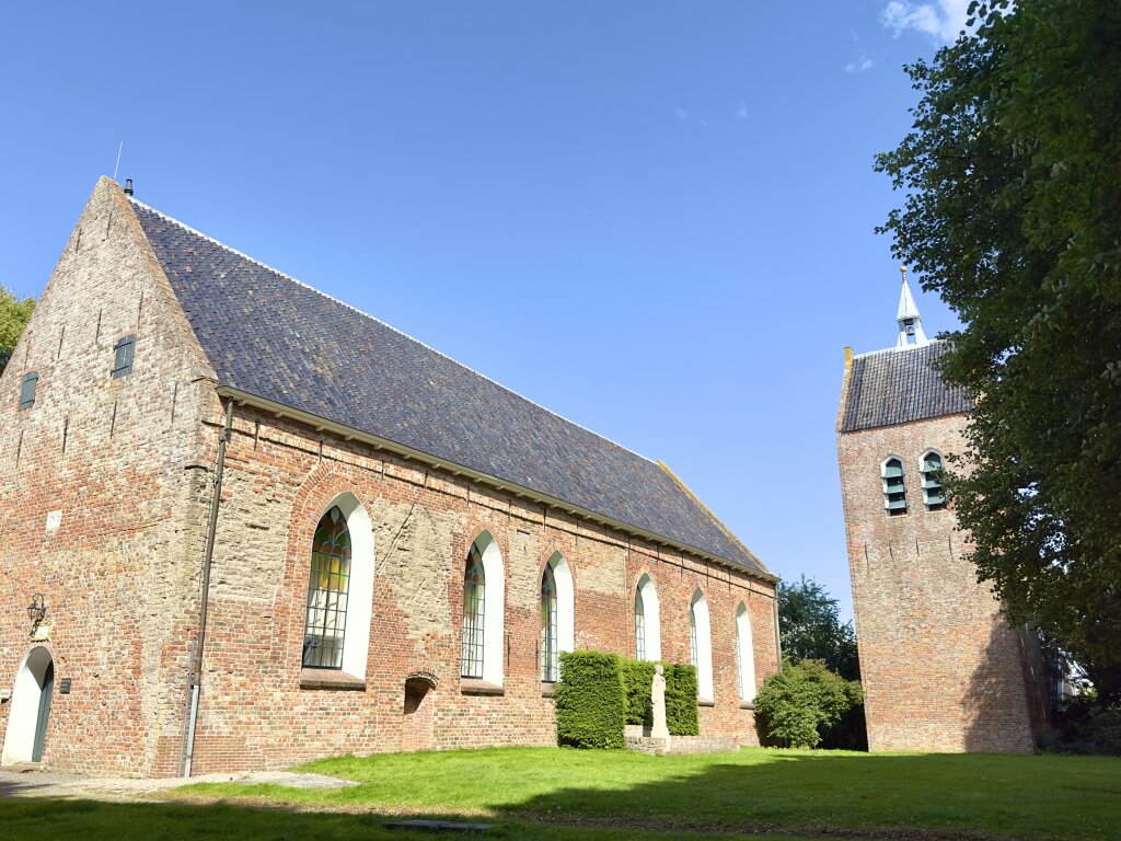 De Laurentiuskerk met toren in Baflo. Omringd door kale bomen, groen gras en een blauwe lucht.